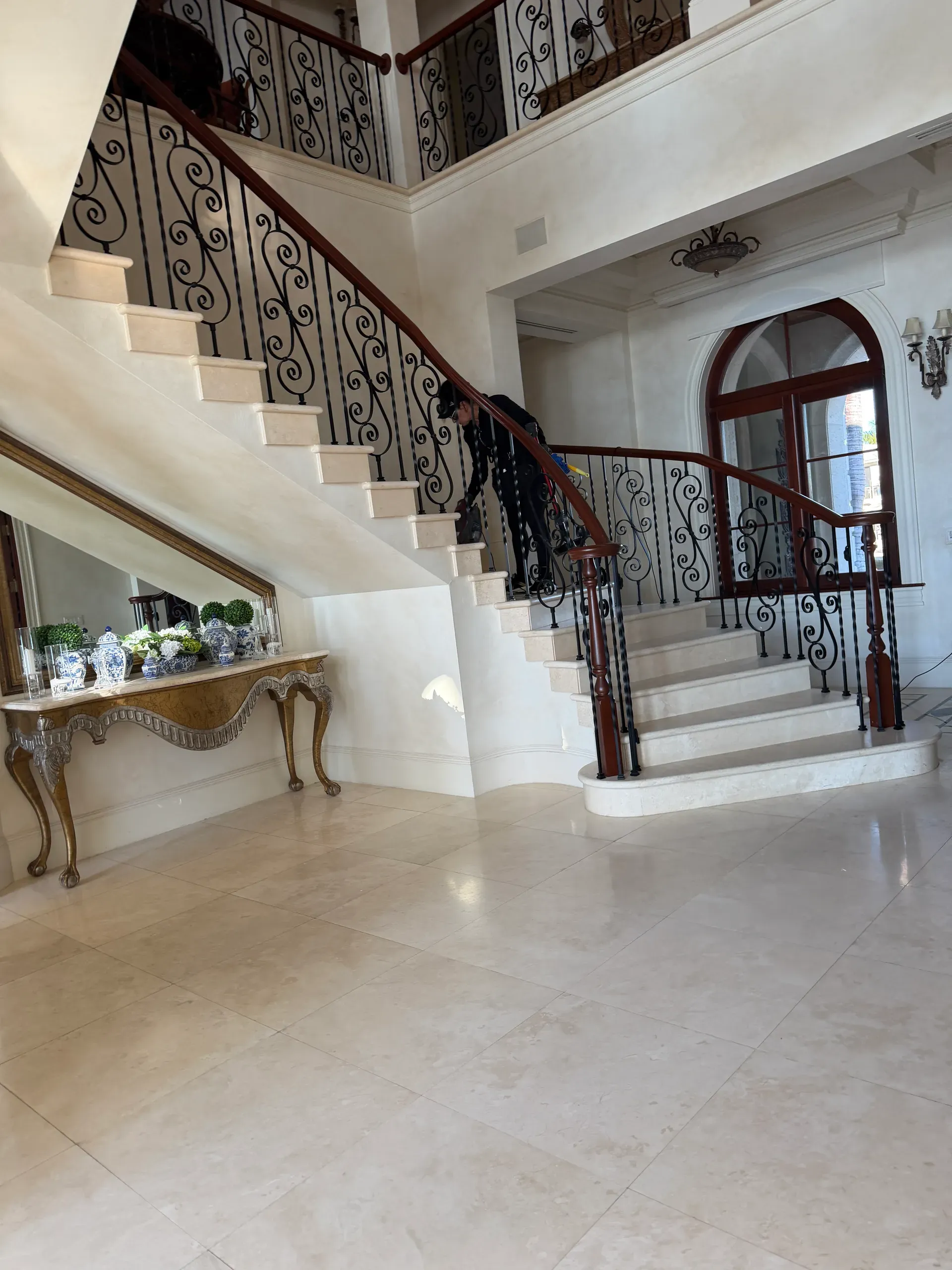 A grand foyer with a curved staircase and wrought-iron railing while a team member cleans the balusters