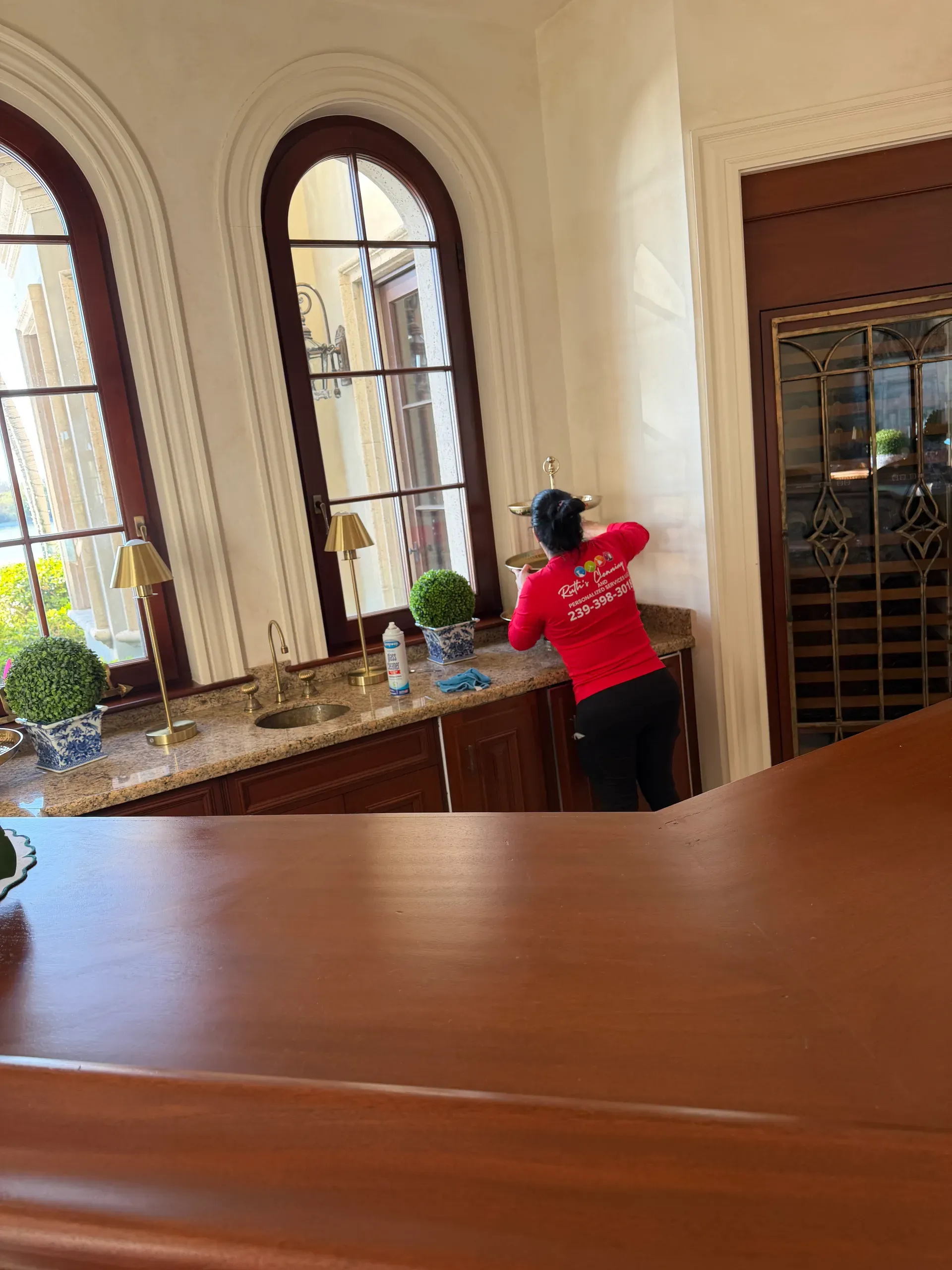 A team member in a red company shirt wiping down a granite wet bar under tall arched windows