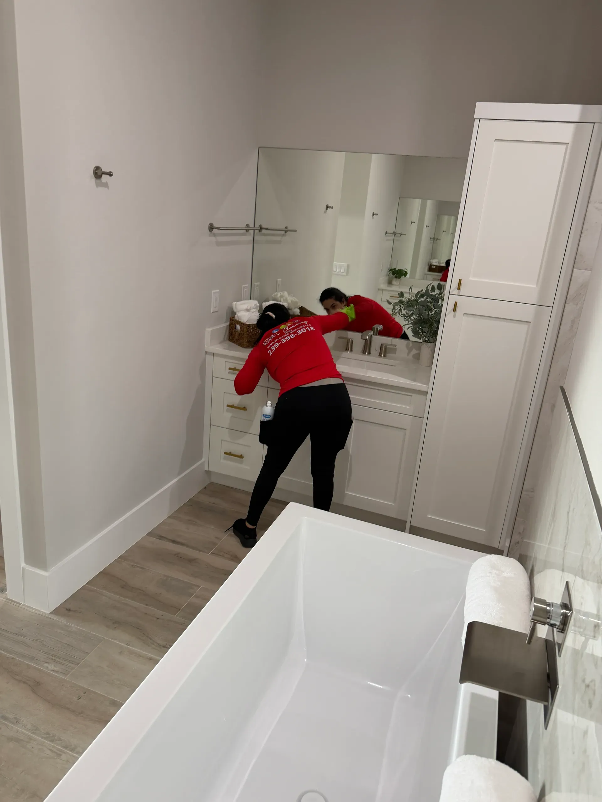 A team member in a red company shirt cleaning a bathroom vanity mirror, reflected in the glass
