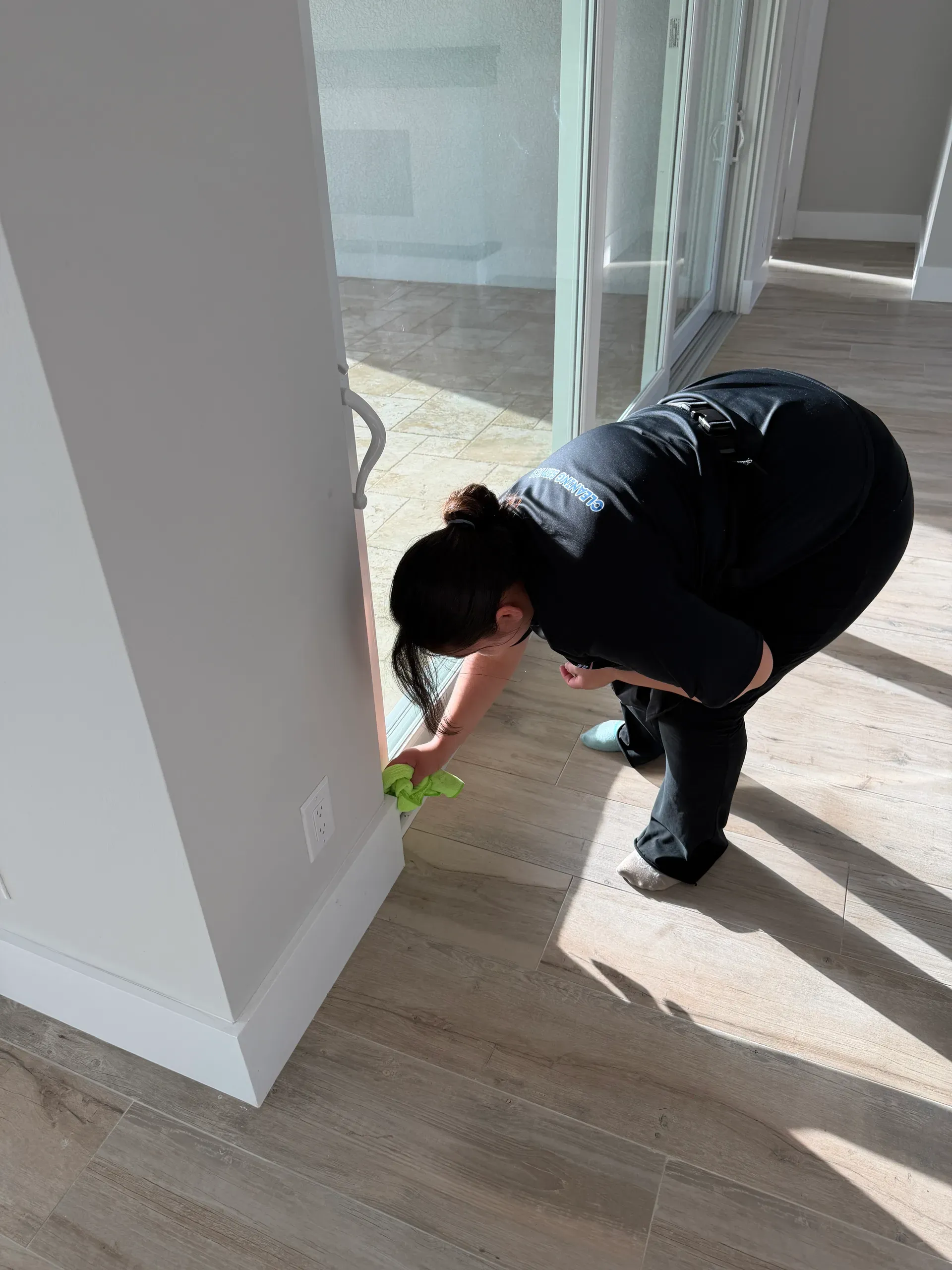 A team member wiping the baseboard beside a sliding glass door in a sunlit room