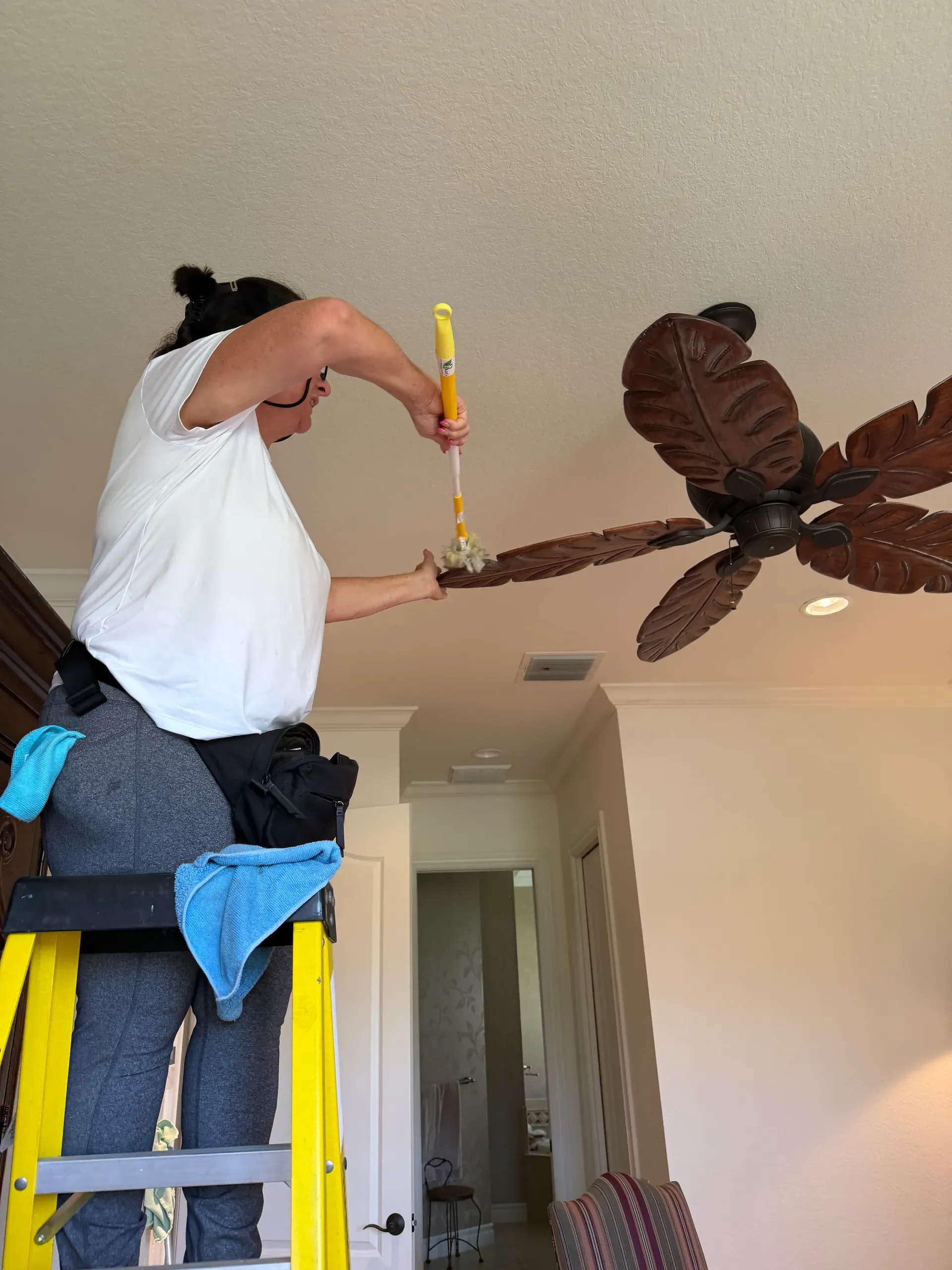 A team member on a yellow ladder dusting a carved wooden ceiling fan blade