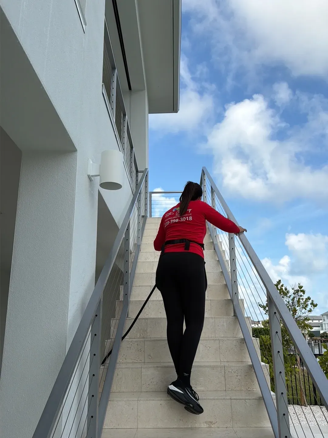A team member in a red company shirt climbing an exterior stone staircase with a hose, under a blue sky