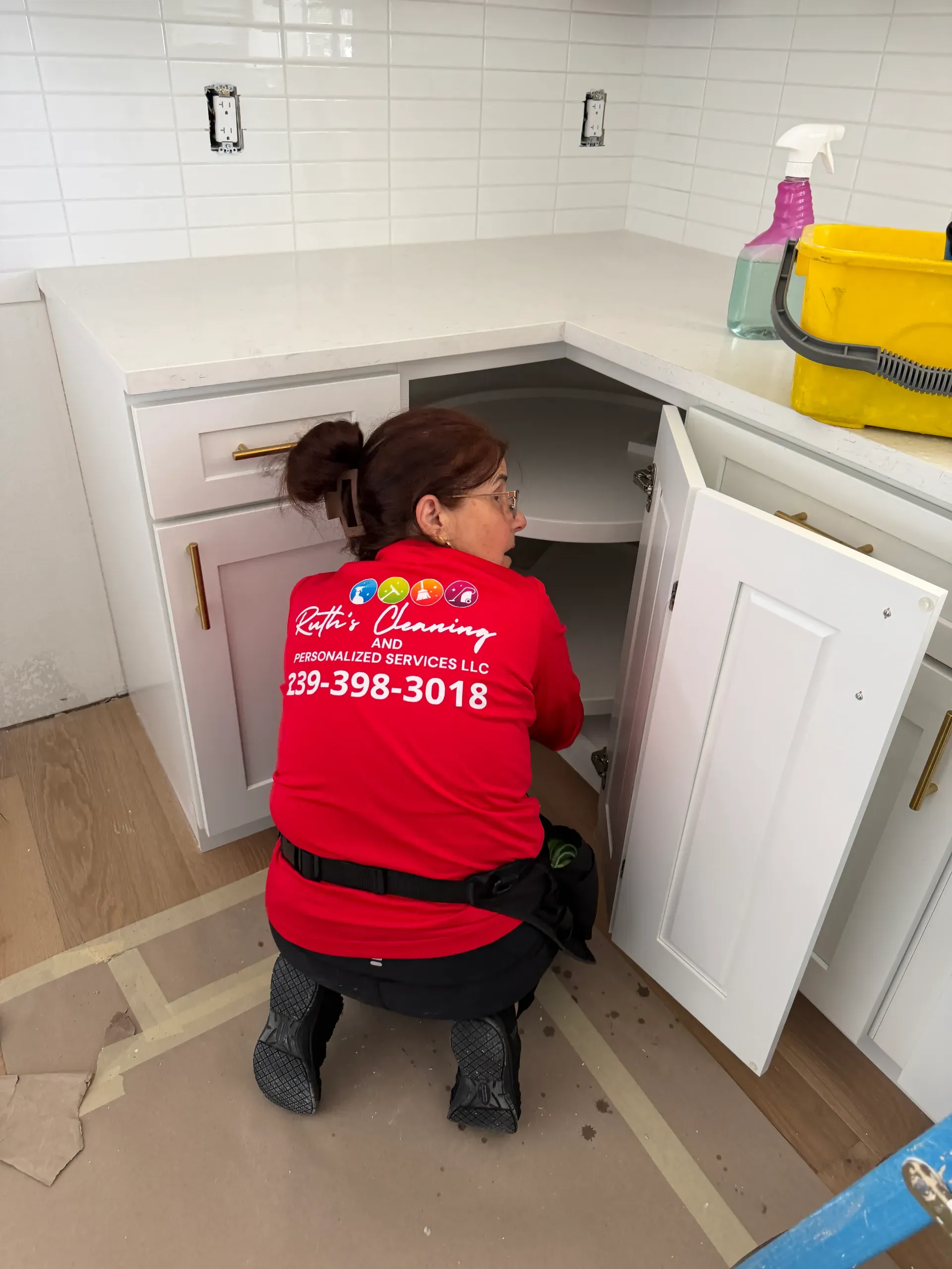 A team member in a red company shirt kneeling to clean inside a corner kitchen cabinet in a newly built kitchen